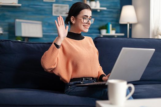 Student waving her friends during online videocall conference sitting on sofa in living room. Young woman using mobile internet web virtual webinar vlass on videoconference meeting