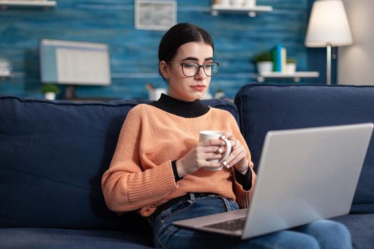 Student reading marketing courses drinking cup of coffee during university seminar. Young woman sitting on sofa in living room checking e-learning platform using laptop computer