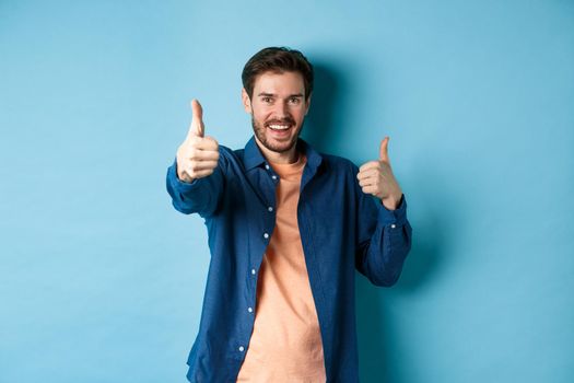Positive smiling guy showing thumbs up and smiling, complimenting you, praising good job, well done gesture, standing on blue background.