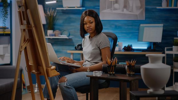 African american artist typing on laptop computer at studio. Black young woman using online technology for drawing creation of modern white vase masterpiece on canvas in art room