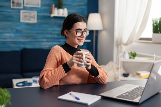 Smiling happy student holding cup of coffee in hands looking at laptop computer sitting at desk. Searching management advertising for university degree browsing e-learning online digital data