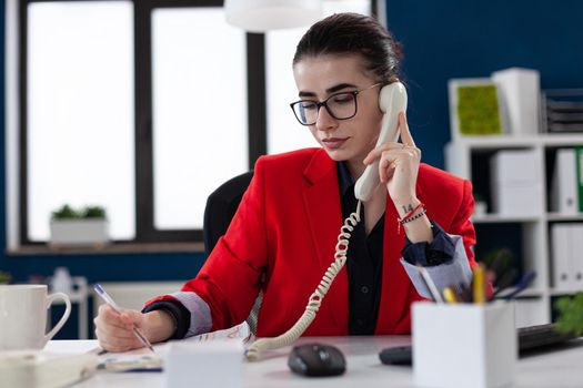 Businesswoman taking notes on clipboard sitting at desk in corporate office, while taking with adviser about financial expertise. Busy manager having a conversation about deadline.