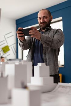 Expert architect looking on smartphone at workplace for designer sketch, building model plan maquette. Professional man working at desk office for construction design and modern structure