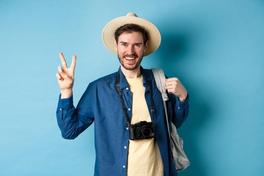 Happy handsome man taking photo on summer vacation, showing peace sign and smiling, wearing straw hat and holding tourist backpack, blue background.