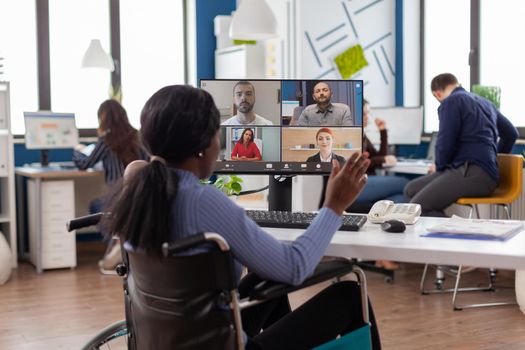 Paralysed handicapped black worker sitting immobilized in wheelchair having videomeeting discussing online with remotely colleagues in business office. Invalid employee working in financial company.