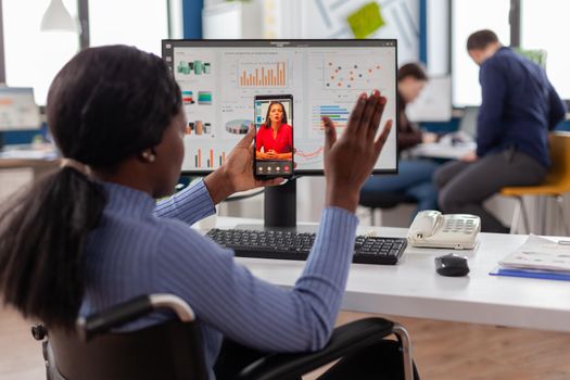 African disabled worker waving at online partner during video call sitting immobilized in wheelchair talking with remote partner on video call, from startup business office discussing project online.