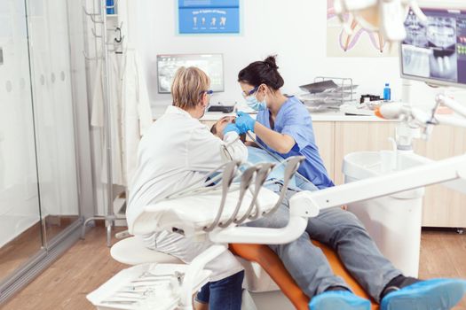 Senior woman stomatologist analyzing teeth of sick patient while sitting on stomatological chair. Doctor speaking to man while nurse cleaning mouth during dental examination in stomatology clinic