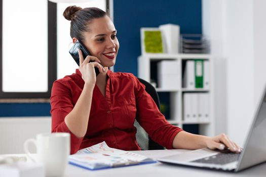 Busy businesswoman answering phone call sitting at desk in corporate workplace , discussing with client. Entrepreneur manager discussing with colleagues during call, looking at laptop screen.