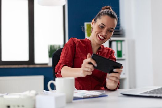 Businesswoman sitting at desk in corporate office playing video games, using smartphone. Entrepeneur taking a break from wwork having fun with games holding phone. Employer having fun at wwork.