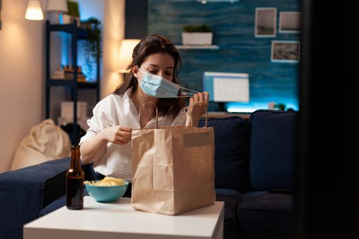 Cheerful woman taking medical protection face mask out after purchasing takeaway food in evening. Smiling caucasian female enjoying delicious junk-food home delivered. Tasting box order