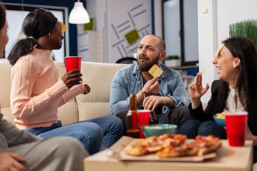 DIverse team of coworkers playing charades after work at office space. Multi ethnic friends enjoy guessing game of imiation as fun cheerful activity entertainment with food and drinks