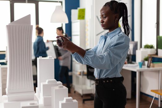 African american woman as professional architect working with smartphone for building model maquette. Engineer looking at modern work concept of construction project design