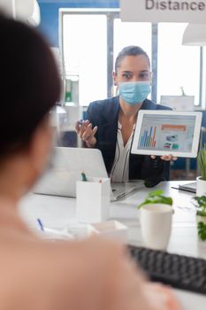 Executive manager with face mask showing statistics to her collegue using digital tablet computer. Coworkers working in new normal office keeping social distance to prevent infection with covid19.