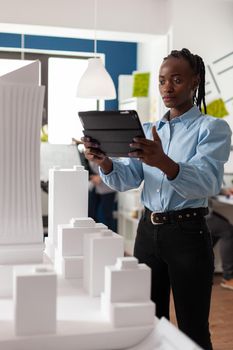 Architect woman of african american ethnicity working on tablet looking at professional maquette building model. Architectural worker viewing design for modern project in development