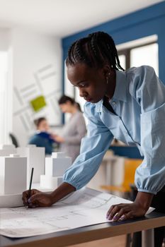 Woman of african american ethnicity working as architect in professional office. Engineer constructor at desk looking on blueprint plan for building model maquette. Development project