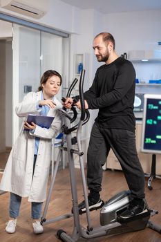 Aathlete running on cross trainer, while medical specialists supervise exercise, controlling physical activity, measuring heart rate in sports science laboratory.