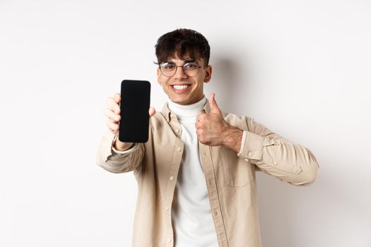 Portrait of handsome young man in glasses, showing empty smartphone screen and thumb up, recommending online app or store, standing pleased on white background.