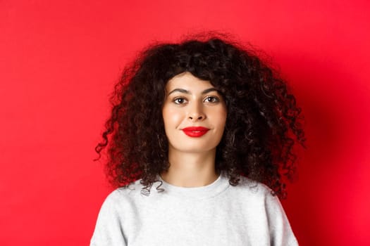 Close up portrait of beautiful modern woman with curly hairstyle and red lips, smiling happy at camera, standing on studio background.