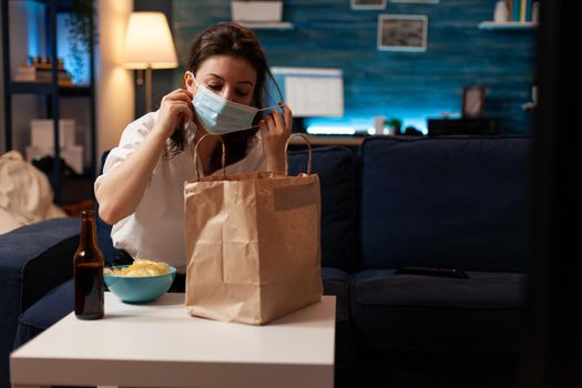 Cheerful woman taking medical protection face mask out after purchasing takeaway food in evening. Smiling caucasian female enjoying delicious junk-food home delivered. Tasting box order