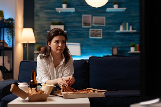 Caucasian female taking tasty delicious pizza slice eating fastfood home-delivered order while watching comedy movie on television. Woman relaxing on couch enjoying takeaway food delivery