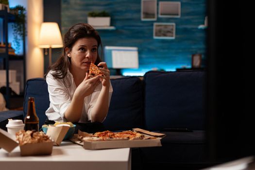 Caucasian female taking tasty delicious pizza slice eating fastfood home-delivered order while watching comedy movie on television. Woman relaxing on couch enjoying takeaway food delivery