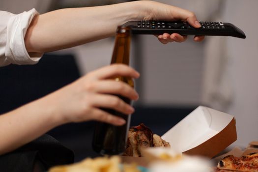 Close-up of woman hands changing channels using remote watching entertainment comedy movie film on television in evening. Caucasian female relaxing having takeaway food home delivered