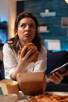 Caucasian female holding tastu burgery in hands browsing on social media using smartphone relaxing on couch during junck-food night. Woman enjoying takeaway food home delivery