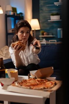 Happy woman changing channels using remote watching entertainment movie on television eating tasty delicious burger in evening. Caucasian female enjoying takeaway food home delivered