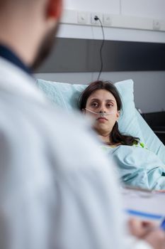 Caucasian female with nasal oxygen tube resting in bed in hospital ward. Practitioner man doctor discussing with patient explaining medical expertise writing test results on clipboard