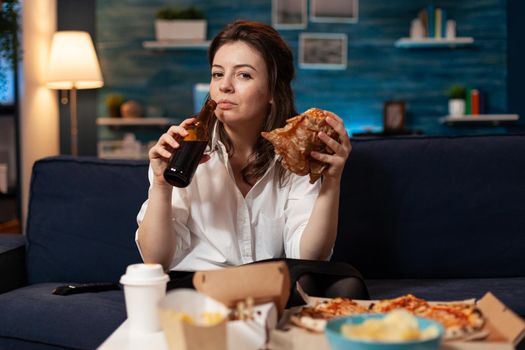 Portrait of woman looking into camera during fastfood lunch meal order relaxing on sofa late at night in living room. Caucasian female enjoying tasty burger, takeaway food home delivered