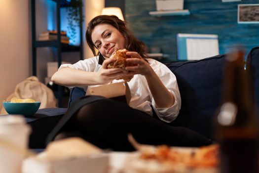 Woman talking at phone with friends while holding tasty delicious burger in hands relaxing on couch in living room late at night. Caucasian female enjoying tajeaway junk food home delivered