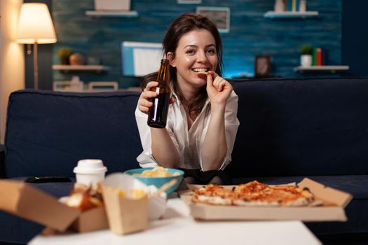 Portrait of caucasian female looking into camera holding beer bottle in hands eating tasty delicious pizza slice during takeaway food home delivered. Woman relaxing on couch late at night