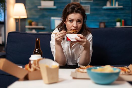 Portrait of caucasian female looking into camera while eating chinese chopstick relaxing on sofa in living room late at night. Woman enjoying takeaway food home delivered. Tasty fastfood lunch order