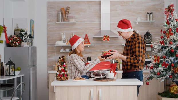 Happy family bringing wrapper christmas present gift with ribbon on it in xmas decorated kitchen celebrating christmas holiday. Grandfather with grandchild wearing santa hat enjoying winter season