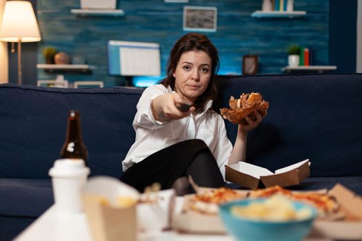 Portrait of woman changing channels using remote watching entertainment movie series sitting on sofa in living room late at night. Caucasian female enjoying takeaway food home delivered