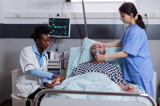 Sick patient lying in bed while woman nurse putting oxygen mask monitoring respiratory illness. Physician black medic writing sickness treatment working in hospital ward during recovery appointment