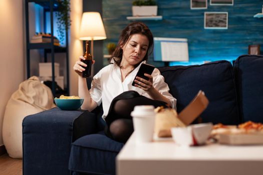 Caucasian female relaxing on couch browsing on social media using smarhphone while drinking beer during relaxing night at home. Woman enjoying takeaway food home delivered. Junk-food order