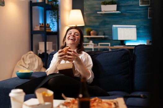 Caucasian female laughting while watching entertainment movie series on television eating tasty delicious fast food burger. Woman enjoying unhealthy takeaway food home delivered. Lunch meal order