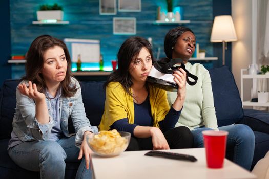 Sad women after losing virtual reality gaming competiton holding vr headset sittingon couch. Mixed race group of people hanging out together having fun late at night in living room