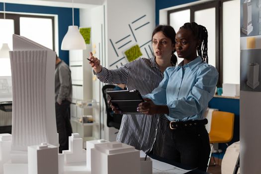 Diverse work team of multi ethnic architects working on tablet checking blueprint plan of building model maquette. Women analyzing project design on paper discussing construction