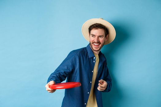 Happy man laughing and playing frisbee, throwing at friend and smiling, standing in summer hat on blue background.