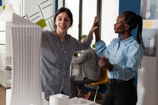 Architecture multi ethnic workmates in partnership to design building model maquette. Diverse professional engineer women highfive after success in blueprint plans cooperation