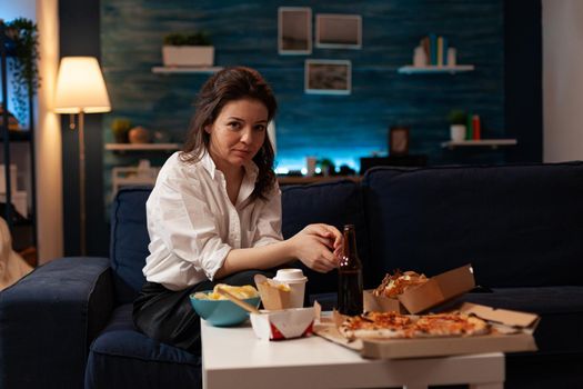 Portrait of smiling woman looking into camera sitting on couch enjoying free time ordering takeaway meal in living room at night. Junk food standing on table. Fast food order. Delivery fast-food box