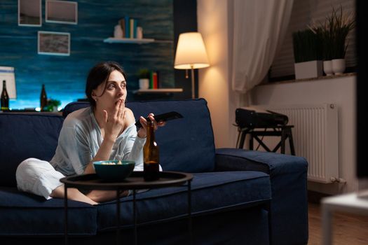 Focused interested woman eating popcorn watching interesting movie show on television. Female sitting on comfortable couch dressed in pajama late at night in living room having concentrated xpression