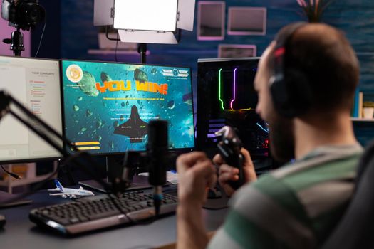 Over shoulder footage of professional streamer playing digital videogames on computer using headphones, microphone and controle. Streaming Man raising hands for wining space shooter game