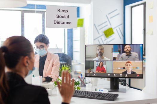 Business woman saying hello during remove video conference in new normal workplace wearing face mask as safety prevetion during global pandemic with covid10. Keeping social distancing from coworker.