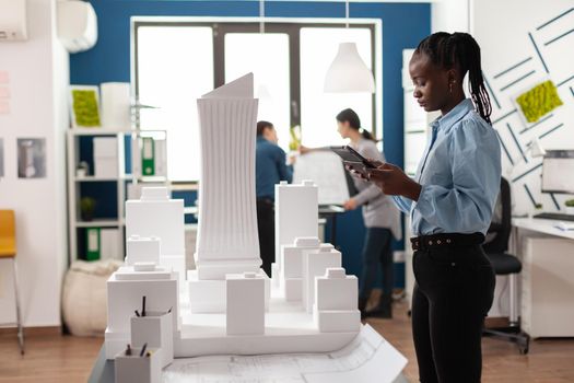 African american architect woman working on tablet looking at building model maquette. Professional engineer with blueprint plans technology viewing modern design concept