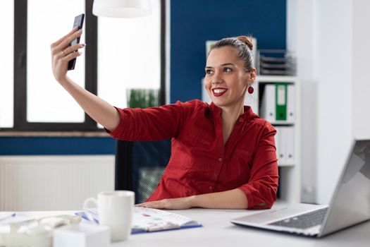 Successful businesswoman having fun at work taking selfies, sitting at desk office workplace using smartphone. Employee using phone camera to take photos of herself during break time.