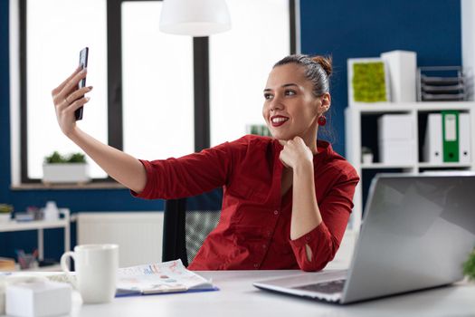 Successful businesswoman having fun at work taking selfies, sitting at desk office workplace using smartphone. Employee using phone camera to take photos of herself during break time.