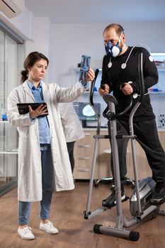 Team of doctor researchers monitoring endurance of man performance sports wearing mask running cross trainer. Lab science doctor measuring vo2 of sportsman, looking at x-ray explaning health status.